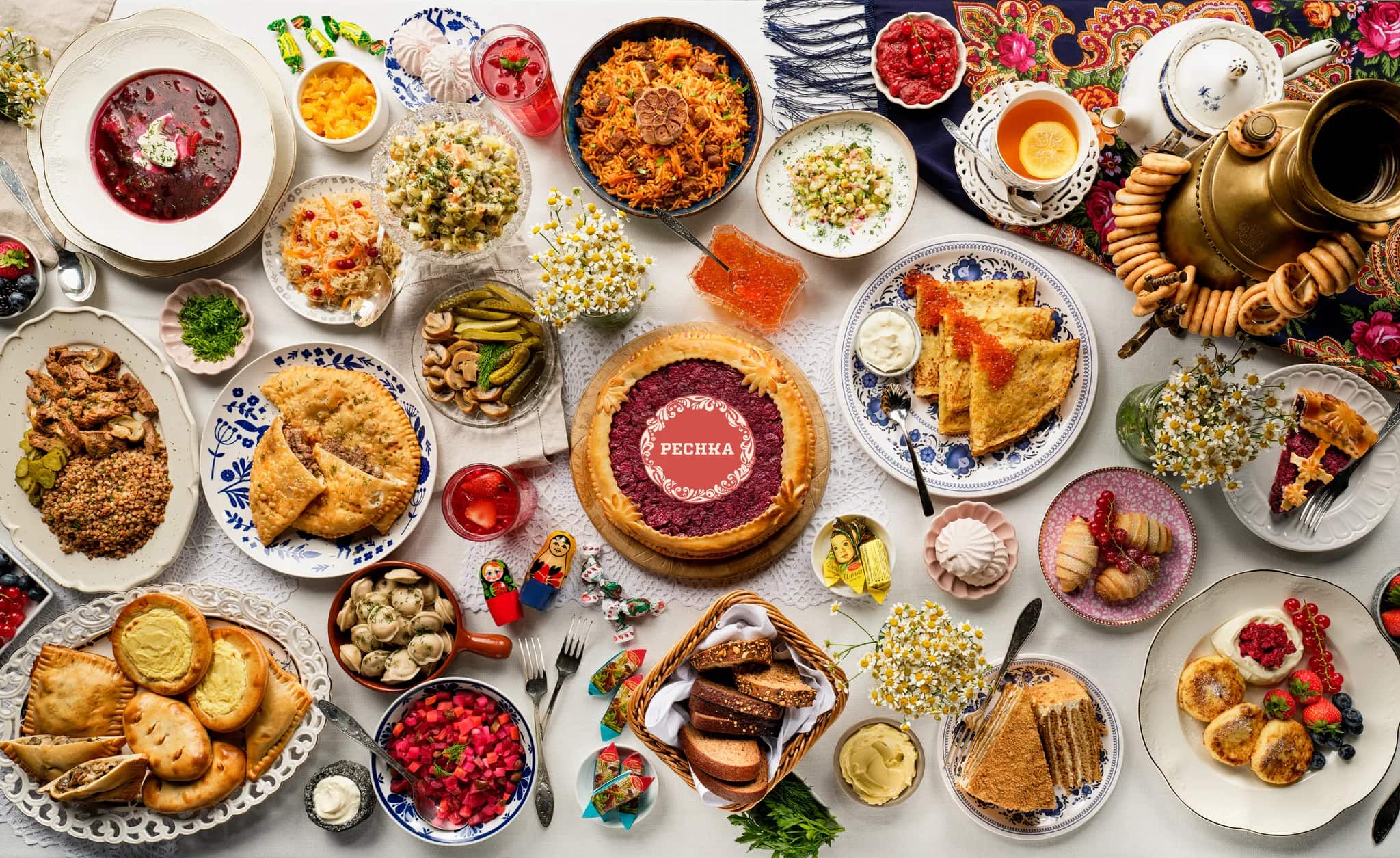 A Pechka table laid with borscht, olivier, plov, pelmeni, blini with caviar, pies and a samovar